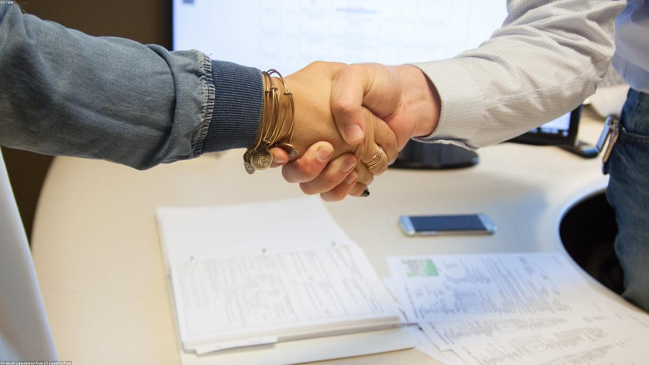 Two people shaking hands across a desk covered with paperwork